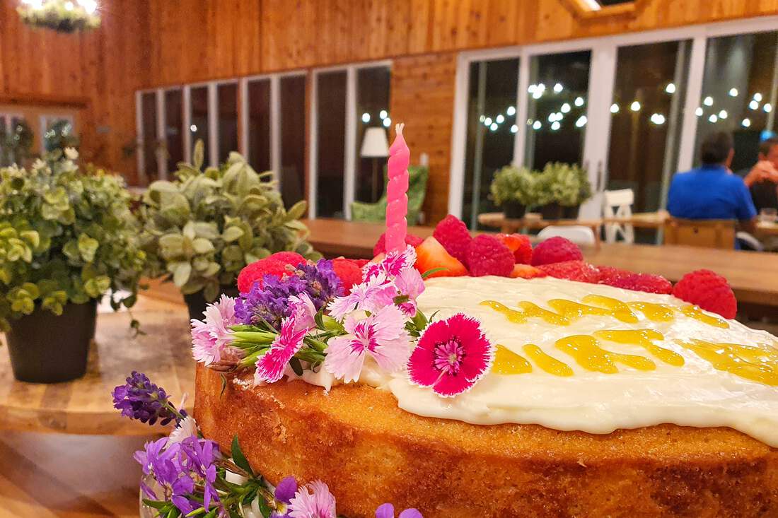 A birthday cake dressed in fresh flowers and berries, the dining hall lit behind it